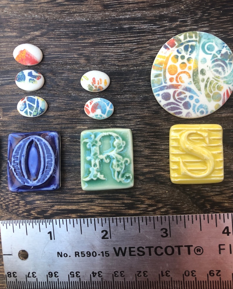 May include: A set of ceramic tiles with colorful designs. The tiles are in the shape of circles, ovals, and squares. The tiles feature a variety of patterns, including floral, geometric, and abstract designs. The tiles are arranged on a wooden surface with a ruler for scale. The ruler is labeled "WESTCOTT" and has markings in inches.