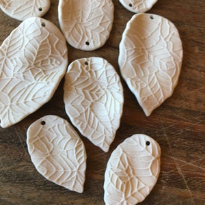 Puede incluir: Una colección de diez colgantes de cerámica blanca en forma de hoja con un delicado patrón en relieve. Cada colgante tiene un agujero en la parte superior para colgar.