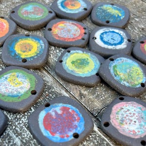 May include: A collection of 14 ceramic tiles with colourful circular designs. Each tile has two holes for hanging. The tiles are black with a variety of colours including yellow, blue, green, red, and white.