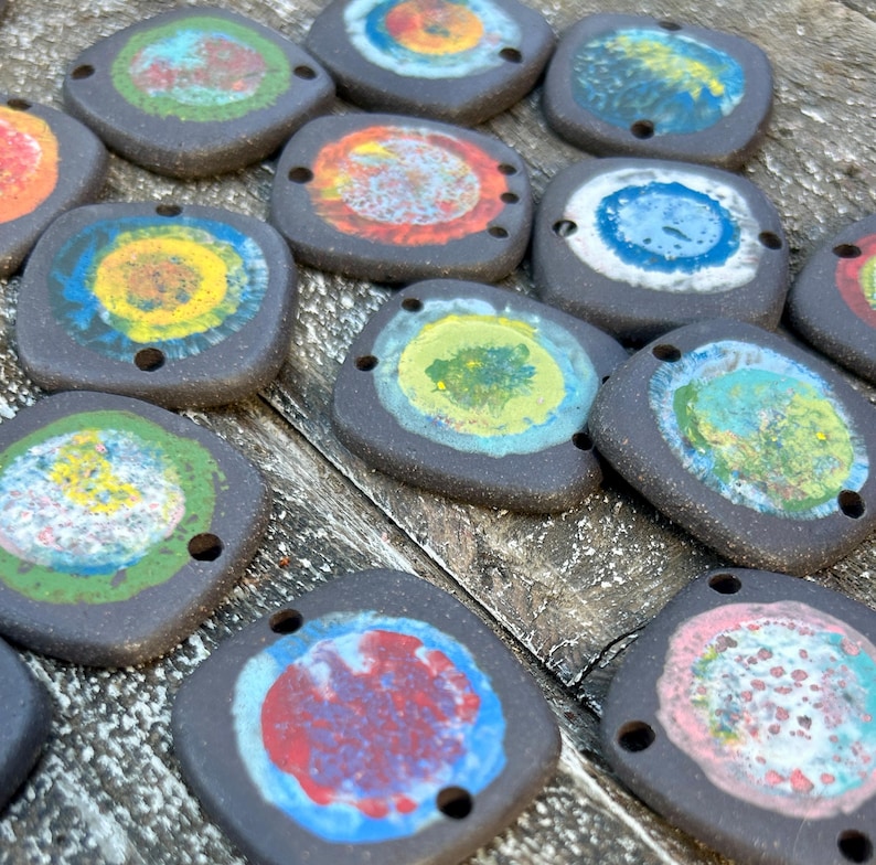 May include: A collection of 14 ceramic tiles with colourful circular designs. Each tile has two holes for hanging. The tiles are black with a variety of colours including yellow, blue, green, red, and white.