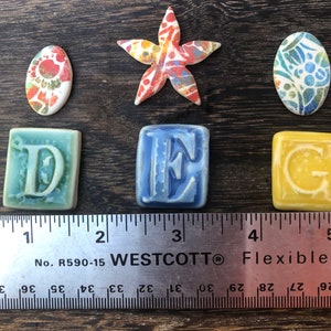May include: A set of ceramic tiles with the letters D, E, and G, along with a starfish and two oval-shaped tiles with colorful patterns. The tiles are arranged on a wooden surface with a ruler measuring 5 inches.