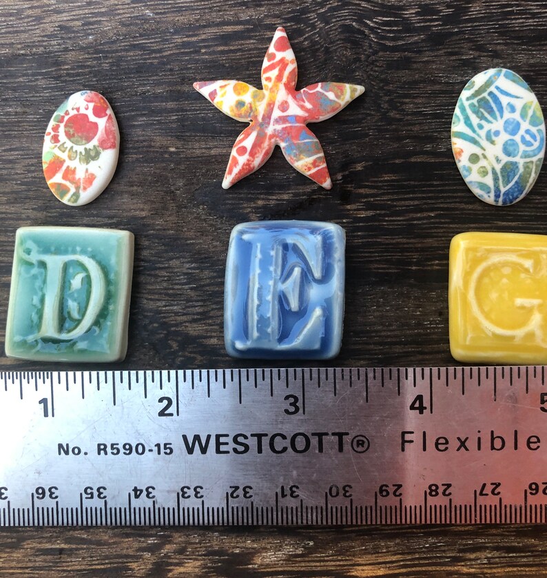 May include: A set of ceramic tiles with the letters D, E, and G, along with a starfish and two oval-shaped tiles with colorful patterns. The tiles are arranged on a wooden surface with a ruler measuring 5 inches.