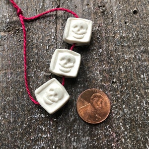 May include: Three white ceramic skull beads strung on a red cord. The beads are square shaped and have a raised skull design.