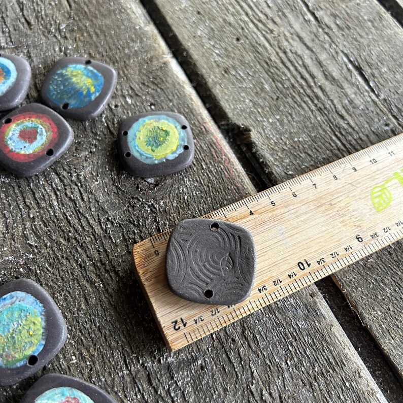 May include: A close-up of a wooden ruler measuring 30 cm, with a black ceramic tile with a swirl design on top. The tile has two holes and is approximately 3.8 cm wide.