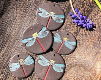 May include: Five round ceramic ornaments featuring a dragonfly design. Each ornament is painted with a blue dragonfly on a dark brown background. The dragonflies have red bodies and black outlines.