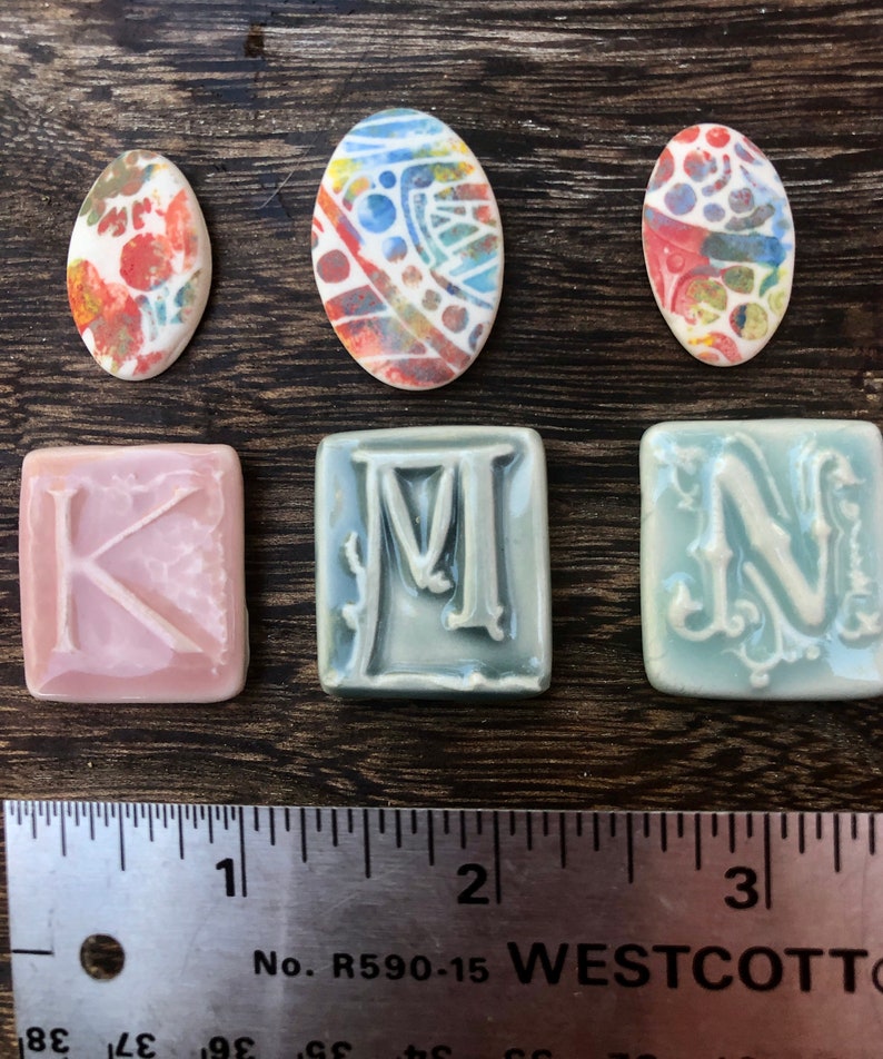 May include: Three ceramic tiles with the letters K, M, and N in a decorative font.  Three oval ceramic tiles with colorful abstract designs.