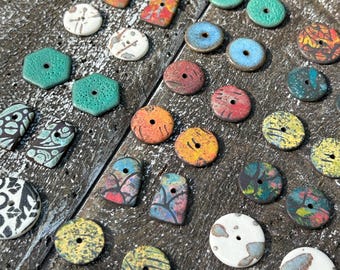 May include: Assortment of colourful ceramic buttons with various patterns and designs. The buttons are arranged on a wooden surface.