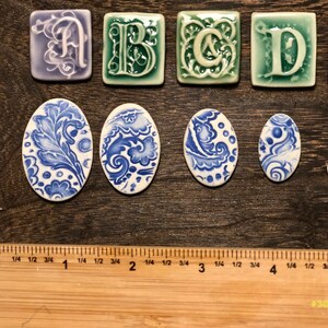 May include: A selection of ceramic tiles, including letters A, B, C, and D in decorative fonts, and oval tiles with blue and white floral designs. The tiles are displayed on a wooden surface, with a ruler for scale.