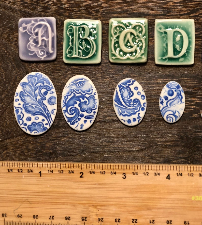 May include: A selection of ceramic tiles, including letters A, B, C, and D in decorative fonts, and oval tiles with blue and white floral designs. The tiles are displayed on a wooden surface, with a ruler for scale.