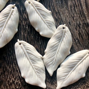 Puede incluir: Cinco adornos de cerámica blanca en forma de hoja con un agujero en la parte superior para colgar. Las hojas tienen una superficie texturizada y un aspecto natural y orgánico.