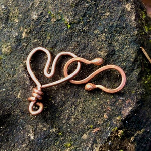 May include: A copper heart-shaped wire charm with a twisted wire detail. The charm is on a rough, brown, textured surface.