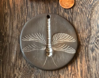 May include: A round, dark brown ceramic ornament featuring a white, detailed dragonfly design. The ornament has a hole at the top for hanging.