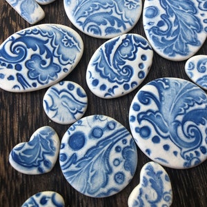 May include: A collection of white ceramic pieces with blue floral patterns. The pieces vary in shape, including ovals, circles, and hearts. The blue designs feature intricate leaf and swirl motifs, creating a classic, decorative aesthetic. The pieces are arranged on a dark wooden surface.