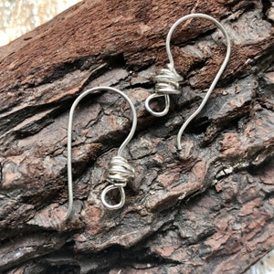 May include: Two silver-colored earring hooks with coiled wire detailing. The hooks are curved and have a small loop at the bottom for attaching charms or beads. The earrings are displayed on a textured, brown wooden surface.