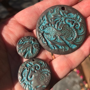 May include: Three round ceramic pendants with a teal blue and brown glaze. The pendants feature an intricate design of a bird with floral accents.