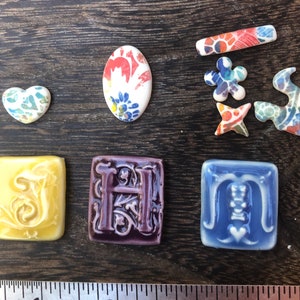 May include: A set of ceramic tiles with various designs, including a heart, an oval, a flower, a star, a swirl, and the letters S, H, and M. The tiles are decorated with colorful floral patterns and have a glazed finish.