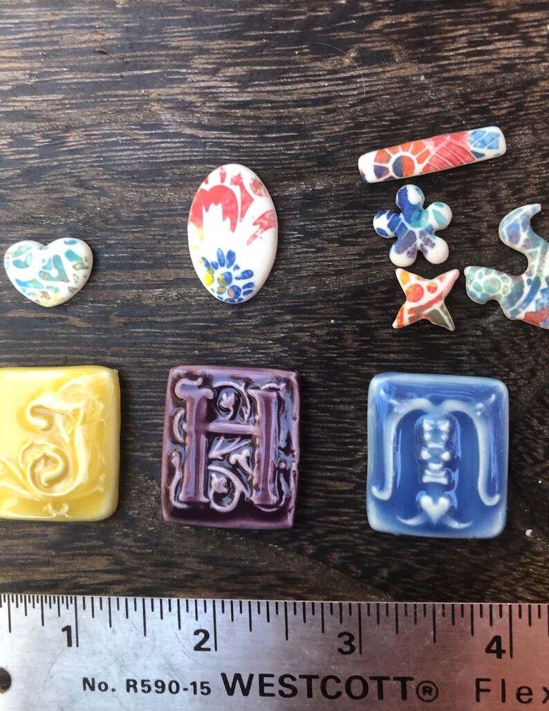 May include: A set of ceramic tiles with various designs, including a heart, an oval, a flower, a star, a swirl, and the letters S, H, and M. The tiles are decorated with colorful floral patterns and have a glazed finish.