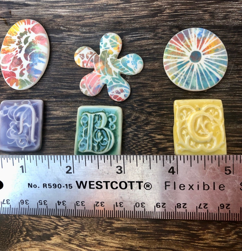 May include: Six ceramic tiles with colorful designs. The tiles feature a flower, a circle with radiating lines, a letter A, a letter B, a letter C, and a swirl pattern. The tiles are arranged in a row on a wooden surface with a ruler below them.