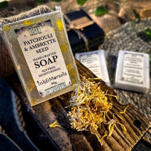 May include: A clear, rectangular bar of hand-crafted soap with yellow floral inclusions, labeled "Patchouli & Ambrette Seed." The label also states the soap is vegan, SLS-free, moisturizing, and compostable. The soap is 90g. Dried yellow flower petals are scattered around the soap.