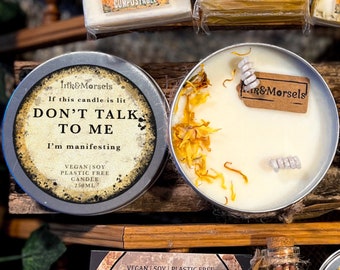 May include: Two round, silver-colored candle tins. One tin has the text "DON'T TALK TO ME, I'm manifesting". The other candle is lit, with dried flowers and a small tag that says "Ink&Morsels". The candles are vegan and soy.