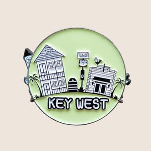 Könnte beinhalten: Ein runder, hellgrüner Emaille-Pin mit einem Key West-Design. Der Pin zeigt Illustrationen von Gebäuden, Palmen und einem Straßenschild mit der Aufschrift „END“. Der Text „KEY WEST“ wird in fetter, weißer Schrift angezeigt.