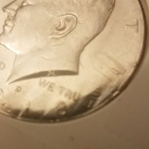 May include: A silver half dollar coin featuring a profile of John F. Kennedy. The coin has the inscription "In God We Trust" and the year "1964".