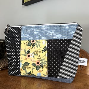 May include: A patchwork zippered pouch with a variety of fabrics. The design includes black polka dots, blue with white dots, yellow floral, black with white crosses, and black and white stripes. A small tag reads "story maker handmade".