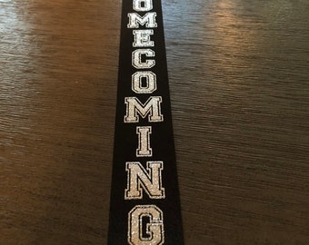 May include: A black ribbon with the word "HOMECOMING" written in silver glitter letters.