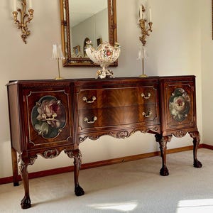 Peut inclure: Un buffet en bois foncé avec des panneaux peints de fleurs et trois tiroirs. Le buffet a une quincaillerie dorée ornée et des pieds en forme de griffes. Un miroir doré et des appliques murales sont montés sur le mur au-dessus du buffet.