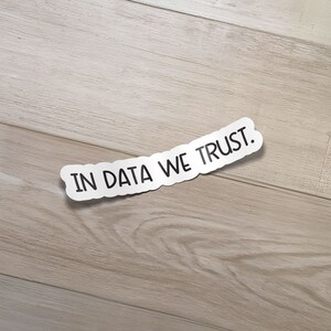 May include: A white sticker with the text "IN DATA WE TRUST." on a light brown wood background.