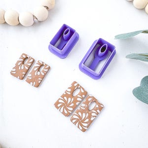 May include: Two purple rectangular clay cutters with integrated oval hole punches, alongside four rectangular clay pieces in a terracotta color featuring a white floral pattern. The clay pieces are arranged in two pairs, each with a pre-cut oval hole.