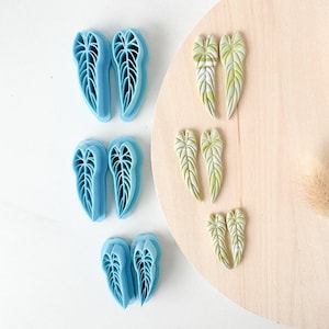 May include: Three pairs of light blue plastic leaf-shaped cutters and three pairs of pale green and white marbled leaf-shaped clay pieces. The cutters feature intricate vein details, and the clay pieces mirror the leaf shape with textured surfaces.