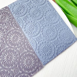 May include: Two textured, rectangular sheets with a floral pattern. One sheet is a muted purple, the other is a light blue. The floral design features large, circular blooms with detailed petal and dot accents. The sheets are likely for crafting or decorative purposes.