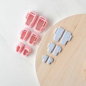 May include: A collection of six tumbler-shaped cookie cutters in varying sizes. Three cutters are light pink and three are light blue. Each cutter features a handle on the side and a tapered body design, arranged in pairs of descending sizes.