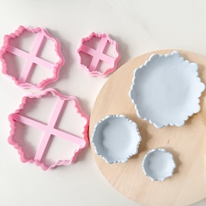 Może przedstawiać: Zestaw różowych foremek do ciastek z falistymi krawędziami w różnych rozmiarach, wraz z trzema jasnoniebieskimi ceramicznymi miseczkami z falistymi krawędziami. Przedmioty ułożone są na jasnej drewnianej powierzchni, tworząc kontrast kolorów i faktur.