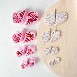 May include: A collection of four pairs of pink plastic seashell-shaped cookie cutters and their corresponding light gray clay or dough impressions. The cutters feature detailed ridged patterns, and the set includes four different sizes arranged in a vertical row.