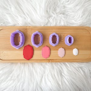 Könnte beinhalten: Ein Set aus vier lila Kunststoff-Ausstechformen in dekorativer Rahmenform, begleitet von vier passenden massiven Rahmen-Ausschnitten in Rot, Pink, Hellrosa und Weiß, angeordnet in absteigender Größe auf einer Holzoberfläche.