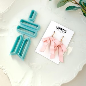 Puede incluir: Pendientes de lazo rosa pálido con aros dorados, presentados en una tarjeta blanca con el texto "Maverly Designs". También se ven cortadores de arcilla polimérica azul claro.