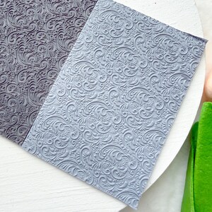 May include: Two rectangular sheets of flexible material featuring a raised, embossed scrollwork pattern. One sheet is dark charcoal gray, and the other is a lighter, pale blue-gray. Both surfaces are covered in intricate, repeating swirls.