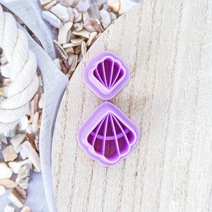 May include: Two purple plastic cookie cutters shaped like seashells. The larger seashell has a more detailed design with ridges and a center line. The smaller seashell has a simpler design.