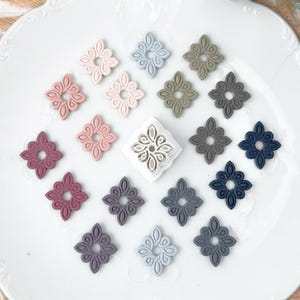 May include: A collection of small, diamond-shaped decorative pieces in various muted colors including pink, blue, green, and gray, arranged around a central white square cutter tool featuring an intricate, symmetrical, leaf-like cutout pattern.