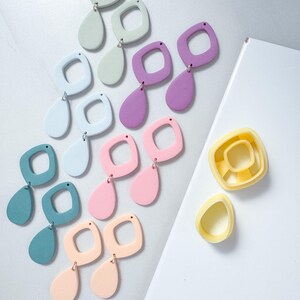 Könnte beinhalten: Eine Sammlung von Hängeohrringen in verschiedenen Pastellfarben wie Minze, Hellblau, Lila, Rosa, Türkis und Pfirsich, jeweils mit einem quadratischen Rahmen und einem tropfenförmigen Anhänger. Rechts liegen zwei gelbe Kunststoffausstecher.