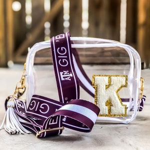 May include: A clear, rectangular crossbody bag with a maroon and white strap. The strap features the text "GIG 'EM" and "AM GIG'E". A gold-colored letter "K" with gold glitter accents is on the front of the bag. A white tassel hangs from the side.