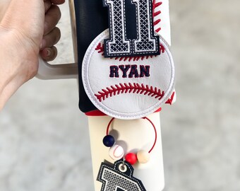 May include: A baseball-themed tumbler wrap with a navy blue and white design. Features a stitched number 11 and a baseball with the name "RYAN". Includes a charm with the letter "R" and a beaded accent.