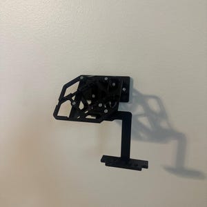 May include: A black wall-mounted display bracket featuring a geometric, open-frame support structure with a flat rectangular base plate and a secondary horizontal shelf component extending outward, secured with visible silver-toned screws.