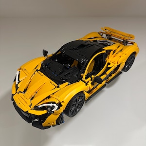 May include: A bright yellow and black model sports car, constructed from interlocking plastic bricks. The car features detailed design elements, including a rear spoiler and black wheels. The model is a replica of a high-performance vehicle.