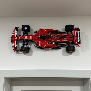 May include: A red Lego Ferrari Formula 1 race car model with black wheels and yellow accents. The car is mounted on a white wall.