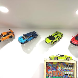 Könnte beinhalten: Fünf Spielzeugautos an einer Wand ausgestellt, darunter ein gelbes und schwarzes Auto, ein blaues und schwarzes Auto, ein grünes und schwarzes Auto, ein rotes und schwarzes Auto und ein orangefarbenes Auto. Die Autos sind an einer weißen Wand mit einer weißen Decke ausgestellt.