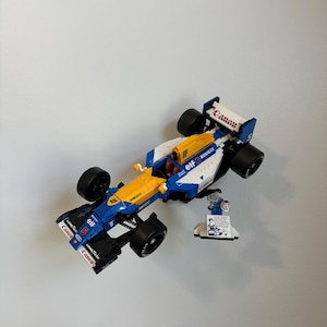 May include: A blue, white, and yellow Lego race car model with black wheels. The model has the Canon, Elf, and Goodyear logos. A small Lego figure and a display stand are next to the car.