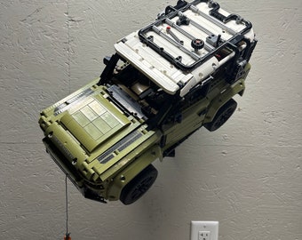 May include: A green Lego Land Rover Defender model with a black roof rack and a white roof. The model is suspended in mid-air with a string attached to the front bumper.